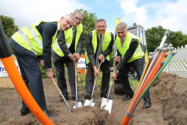 4 Herren im Anzug und Sicherheitsweste auf einer Baustelle beim feierlichen Spatenstich inkl. Glasfaserkabeln im Vordergrund